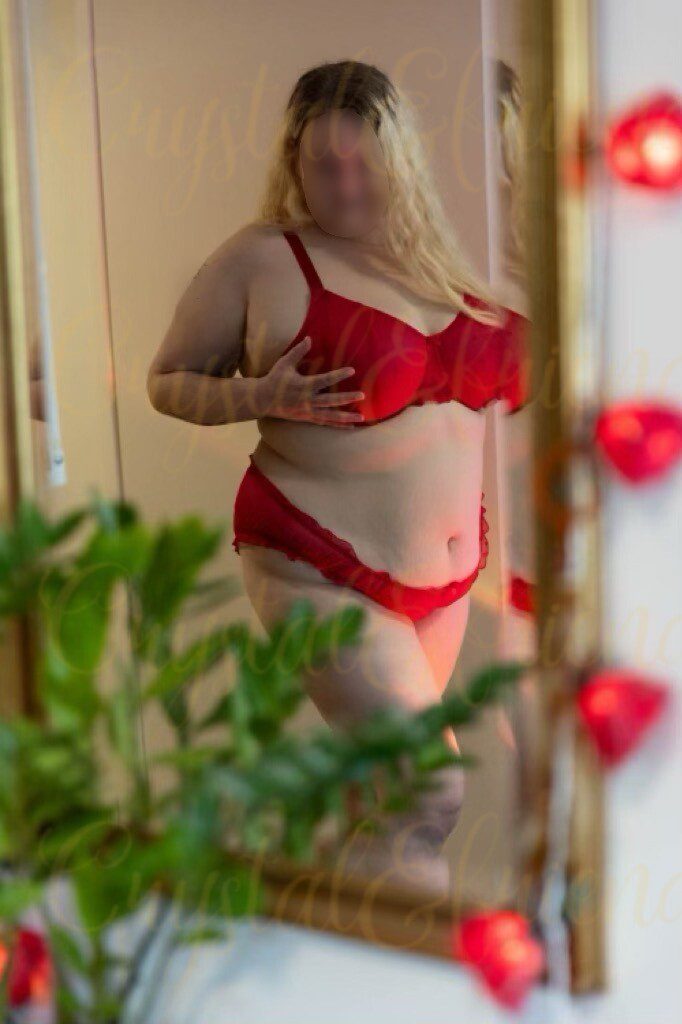 Curvy blond erotic masseuse in red lingerie standing in front of a mirror